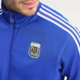 Campera deportiva Adidas Originals de la Selección Argentina, color azul con las icónicas tres tiras blancas en las mangas. Presenta cuello alto, cierre frontal completo, logo del trébol de Adidas en el pecho derecho y escudo de la AFA en el pecho izquierdo.