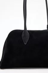 Cartera tipo bowling de diseño estructurado en color negro, con acabado aterciopelado y doble asa larga para llevar al hombro. Presenta un detalle de pequeña tira con broche metálico en la base de una de las asas.