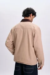 Campera color taupe de corte recto, con cuello camisero de pana marrón oscuro, cierre frontal oculto con botones a presión y grandes bolsillos frontales con solapa.