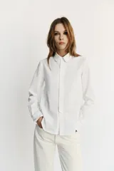 Camisa blanca de manga larga con cuello redondo tipo bebé y cierre frontal de botonadura simple.
