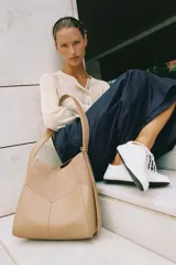 Bolso de hombro color tostado con diseño estructurado que combina paneles de cuero liso y gamuza con pespuntes visibles. Cuenta con un asa larga de hombro ajustable y cierre superior con botón imantado.