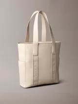 Cartera tote Calvin Klein de color beige, confeccionada en suave piel sintética, con diseño estructurado, asas fijas y compartimentos exteriores en los laterales. Presenta detalles de cinta con logo en jacquard y logo metálico grabado en la parte delantera.