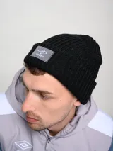 Gorro tipo beanie tejido en punto acanalado, color negro, con parche frontal rectangular gris que presenta el logo de Umbro bordado.