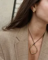 Collar con dije colgante de forma orgánica e irregular, acabado dorado, suspendido de un cordón negro de algodón natural.