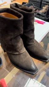 Botas de caña media confeccionadas en cuero gamuzado color marrón oscuro. Presentan un diseño versátil 2 en 1 que permite usar la caña extendida o doblada, con puntera cuadrada y taco bajo.