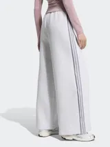 Pantalón deportivo Adidas modelo Spacer, color gris melange, de corte holgado y pierna ancha con pliegues frontales. Cuenta con cintura elástica, bolsillos laterales y las clásicas tiras laterales de la marca en tono gris oscuro, además del logo del Trifolio bordado.