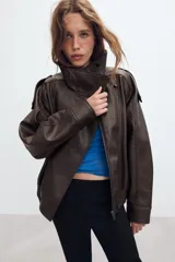 Cazadora tipo biker de cuero sintético en color marrón oscuro, con cuello subido, cierre frontal cruzado con botones forrados y cremallera oculta. Presenta mangas largas con trabillas en los hombros, bolsillos laterales de vivo y bajo con acabado elástico.