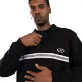 Buzo negro con cuello polo y cierre de un cuarto. Presenta una franja horizontal blanca con texto negro en el pecho y un logo ovalado blanco en el lado derecho. Tiene bolsillos laterales y puños y dobladillo acanalados.