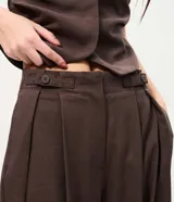 Pantalón sastrero de corte amplio y tiro alto, color marrón, con pinzas frontales profundas y cierre lateral con botón en la pretina.