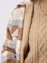 Sobrecamisa de estilo leñador con estampado de cuadros en tonos marrones, beige y gris. Presenta cuello clásico, cierre frontal con botones, bolsillos delanteros y puños tejidos acanalados. El interior está forrado con tejido tipo sherpa para mayor abrigo.