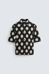 Camisa de manga corta con cuello solapa y cierre frontal de botones, confeccionada en tejido de crochet con patrón de flores en negro y beige.