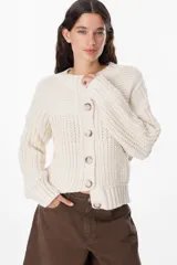 Saco de punto grueso con textura acanalada, cuello redondo y cierre frontal con botones grandes. Presenta un diseño de calce relajado con mangas largas y puños ajustados.