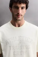 Camiseta de corte regular fit confeccionada en algodón, con cuello redondo y manga corta. Presenta un estampado frontal con el texto 'ATHLETICZ TRAINING CREW' con efecto desgastado.