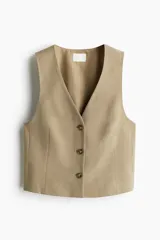 Chaleco corto de color beige, con escote en V pronunciado y cierre frontal de tres botones. Presenta un diseño entallado con pinzas moldeadoras en la parte delantera y está completamente forrado.