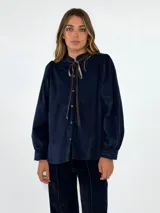 Camisa de pana azul marino con cuello alto, volados en el escote y lazo decorativo. Presenta cierre frontal con botones y mangas largas con puños ajustados.