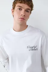 Camiseta de manga larga color blanco, con cuello redondo y corte holgado. Presenta un estampado tipográfico en el pecho y detalles gráficos en la manga.