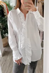 Camisa blanca de algodón con cuello clásico, cierre frontal con botones, mangas largas con puños abotonados y volados en el frente y los puños.