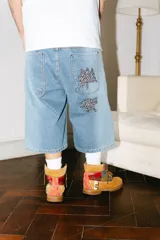 Bermuda de jean de corte baggy, lavado celeste con efecto desgastado. Presenta bordados grandes en los bolsillos traseros con caracteres chinos en negro y blanco. Tiene un parche de cuero marrón en la cintura trasera.