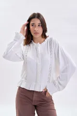 Blusa blanca de manga larga con detalles de encaje vertical en el frente, cuello con volados y terminación con volado en el ruedo. Presenta puños con volados y cierre frontal con botones.