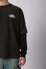 Remera de doble capa con diseño de efecto superpuesto. La capa exterior es de algodón con lavado acid wash en color gris oscuro y cuenta con un logo ovalado estampado en el pecho. La capa interior es de manga larga en color negro liso. Presenta un corte relajado y cuello redondo.