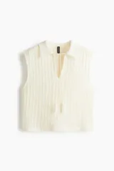 Chaleco de punto color blanco roto, sin mangas, con cuello polo y abertura en V con cordones terminados en borlas.