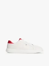 Zapatillas Tommy Hilfiger de diseño clásico y bajo, color blanco con detalles en rojo en la parte trasera del talón y en la lengüeta. Presentan cierre de cordones y el logo metálico distintivo de la marca en el lateral.