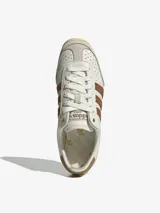 Championes Adidas modelo Japan, con diseño retro de perfil bajo. Presentan un cuerpo de cuero color crema con perforaciones en la puntera, icónicas tres tiras laterales en color marrón y refuerzos de gamuza gris en la puntera y el talón. Incluye suela de goma color amarillento y detalles dorados en el lateral y la lengüeta.