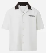 Camisa de manga corta, corte boxy, color crema, con cuello inglés y botones frontales. Presenta un bordado pequeño en el pecho izquierdo con texto en color marrón oscuro.