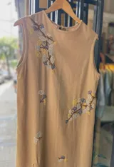 Vestido midi sin mangas color tostado, confeccionado en lino de algodón, con delicados bordados artesanales a mano de ramas con hojas en tonos blancos, azules y amarillos.