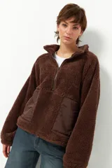 Buzo de tela tipo teddy color marrón, con cuello alto y cierre frontal parcial. Presenta bolsillos frontales en tela de nylon a tono y diseño de manga larga.