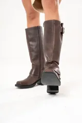 Bota biker de caña alta color marrón, confeccionada en cuero. Presenta hebilla en la parte superior, detalle de aro en la parte inferior y cierre lateral.