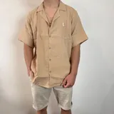 Camisa de hombre color verde oliva, de muselina con textura arrugada, manga corta, corte oversize, cuello cubano y cierre con botones. Presenta un pequeño logo bordado en el pecho.