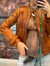 Chaqueta biker corta confeccionada en 100% cuero genuino ovino, color camel. Cuenta con cuello mao con tira y hebilla metálica, bolsillos frontales con cierre, detalles de trabillas con botones y tiras ajustables en los puños.