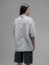 Camisa de manga corta color beige con cuello clásico y cierre de botones. Presenta un diseño de bordado calado con motivos florales y geométricos.