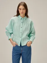 Camisa de lino color verde agua, con cuello clásico, cierre frontal de botones y mangas largas con puños. Presenta detalles de costuras en zigzag en la zona de los hombros y canesú.