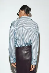 Camisa de corte oversize con estampado de rayas verticales en tonos celeste y negro. Presenta cuello solapa, cierre frontal con botones y mangas largas con puños rectos.