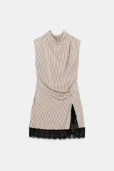 Vestido corto de cuello subido y sin mangas, con diseño fluido y pliegues en los hombros. Presenta un detalle de frunces en el frente, una abertura lateral y un borde inferior decorado con encaje negro en contraste.