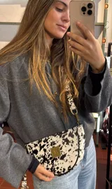 Bandolera con silueta de media luna y diseño estructurado. Confeccionada en cuero con estampado animal print tipo vaca, cuenta con cierre superior, herrajes metálicos y placa frontal dorada con logo. Incluye correa regulable para llevar al hombro o cruzada.