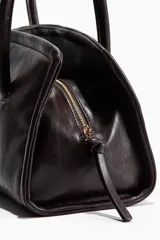 Cartera de hombro rectangular con acabado brillante, cierre superior y asas largas unidas por una trabilla con hebilla metálica.