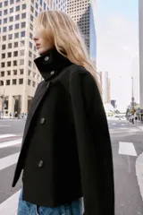 Abrigo corto de paño en color negro, con cuello subido, cierre frontal cruzado con botones, mangas largas con trabillas en los puños y bolsillos delanteros de vivo.