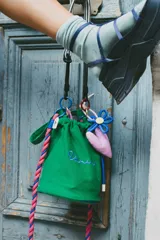 Cartera tipo bucket verde con base estructurada, cordón de ajuste rosa y azul, y llaveros colgantes de flor azul y corazón rosa.