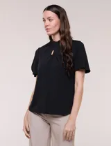 Blusa blanca de corte recto con cuello alto torzado, abertura frontal en gota y mangas cortas con aberturas sobre los hombros.