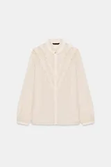 Camisa de cuello redondo y manga larga con puños. Presenta un diseño semitransparente con textura de pequeños puntos en relieve y volantes decorativos en el frente. Cierre frontal con botones ocultos.