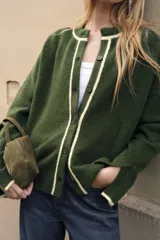 Chaqueta de punto color verde oscuro con cuello redondo y cierre frontal de botones. Presenta detalles de vivos en color crema en el cuello, la tapeta de botones y los puños.