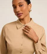 Vestido corto color beige, estilo chemise, confeccionado en tricolina. Tiene cuello inglés, mangas largas con puños abotonados, abotonadura frontal y falda con pliegues.