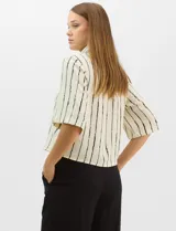 Camisa de corte holgado y silueta recta, con estampado de rayas verticales irregulares en color negro sobre fondo crema. Presenta cuello camisero clásico, mangas cortas amplias y cierre frontal con botones.