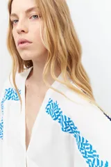 Túnica blanca de lino con corte oversized, escote en V, cierre con botones y mangas cortas. Presenta bordados geométricos azules en hombros y laterales.