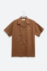 Camisa de hombre color marrón claro, de calce relajado y corte cuadrado, confeccionada en lino y algodón. Tiene cuello tipo camp, cierre con botones de coco grabados con el logo y bordado sutil de flores en el pecho izquierdo.