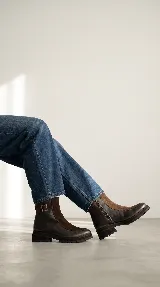 Bota urbana de caña media, confeccionada en una combinación de cuero gastado y gamuza. Presenta una solapa frontal en gamuza, cierre lateral y una tira decorativa con hebilla metálica en el tobillo. Cuenta con una suela de goma robusta y dentada.