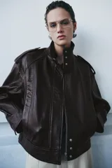 Chaqueta tipo bomber de piel 100% color bordó, con cuello alto, manga larga con trabillas y hombros con trabillas. Tiene bolsillos delanteros y cierre frontal con cremallera y botones ocultos por solapa.